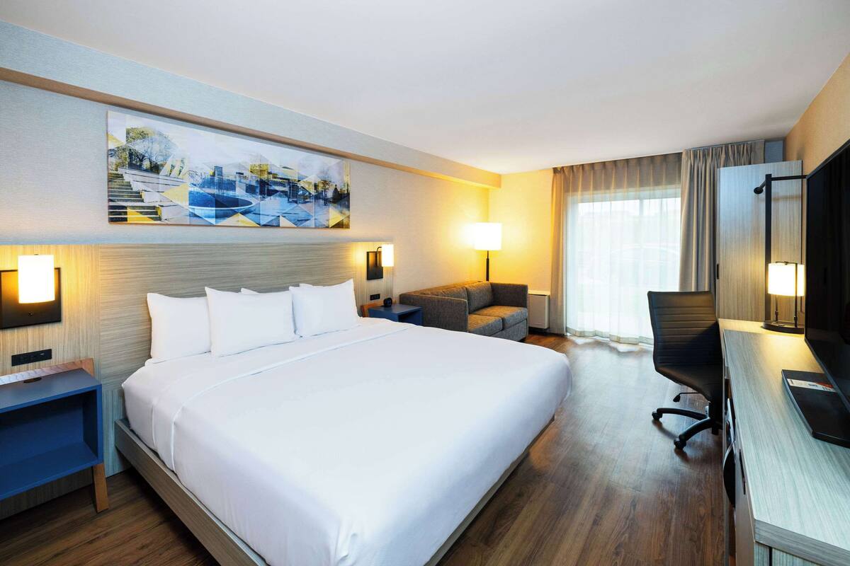 A hotel room with a large bed, a desk, a chair, a sofa, a TV, and a painting on the wall.