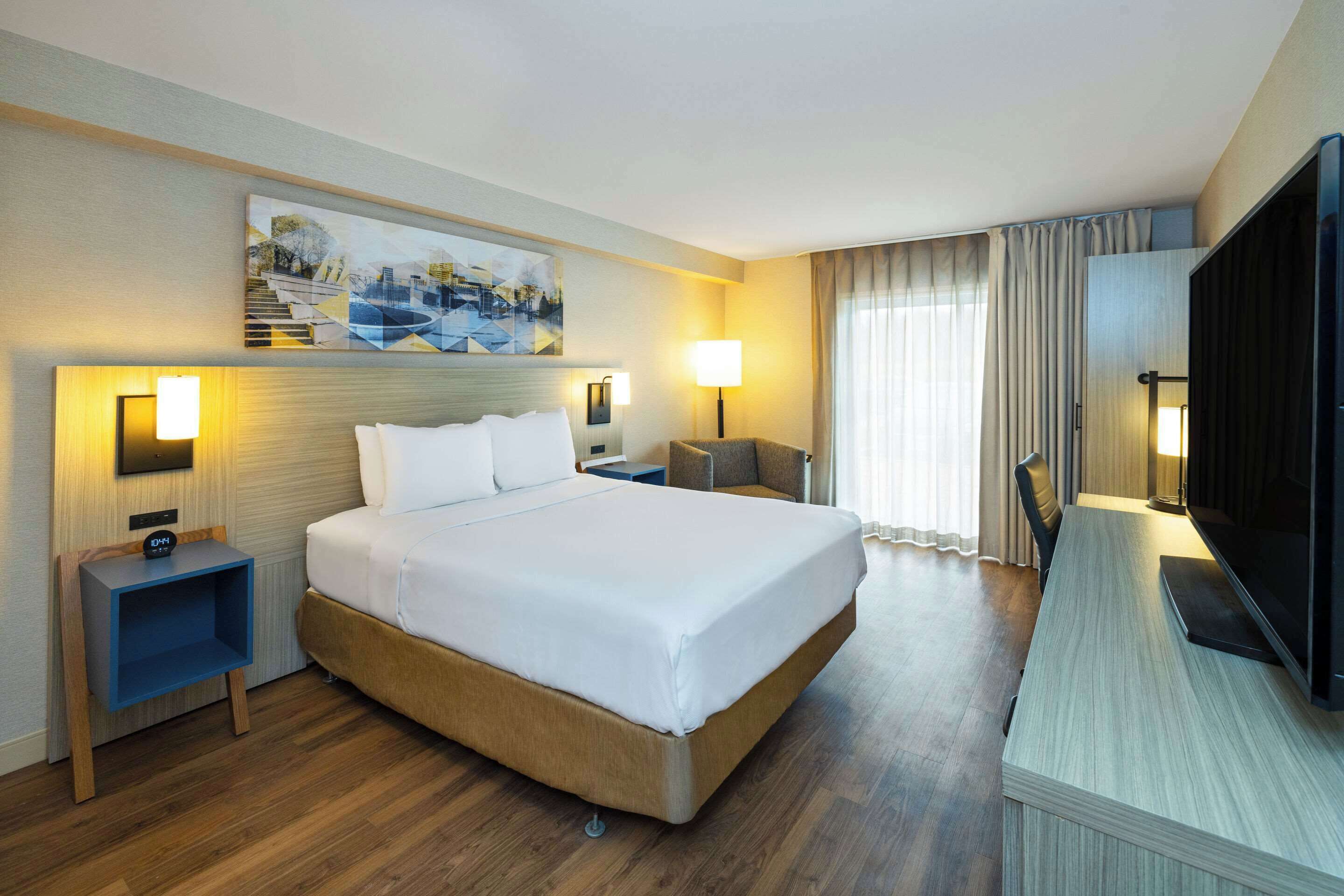 Een hotelkamer met een groot bed, nachtkastjes, een bureau met stoel, een televisie en een schilderij aan de muur.