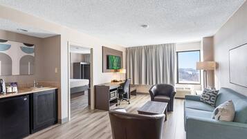 A hotel room with a kitchenette, a dining area, a living room with a sofa and armchair, a flat-screen TV mounted on the wall, and a large window with curtains.