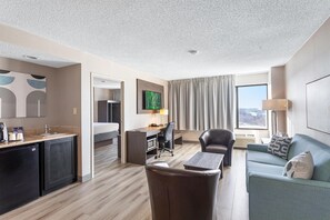 A hotel room with a kitchenette, a dining area, a living room with a sofa and armchair, a flat-screen TV mounted on the wall, and a large window with curtains.