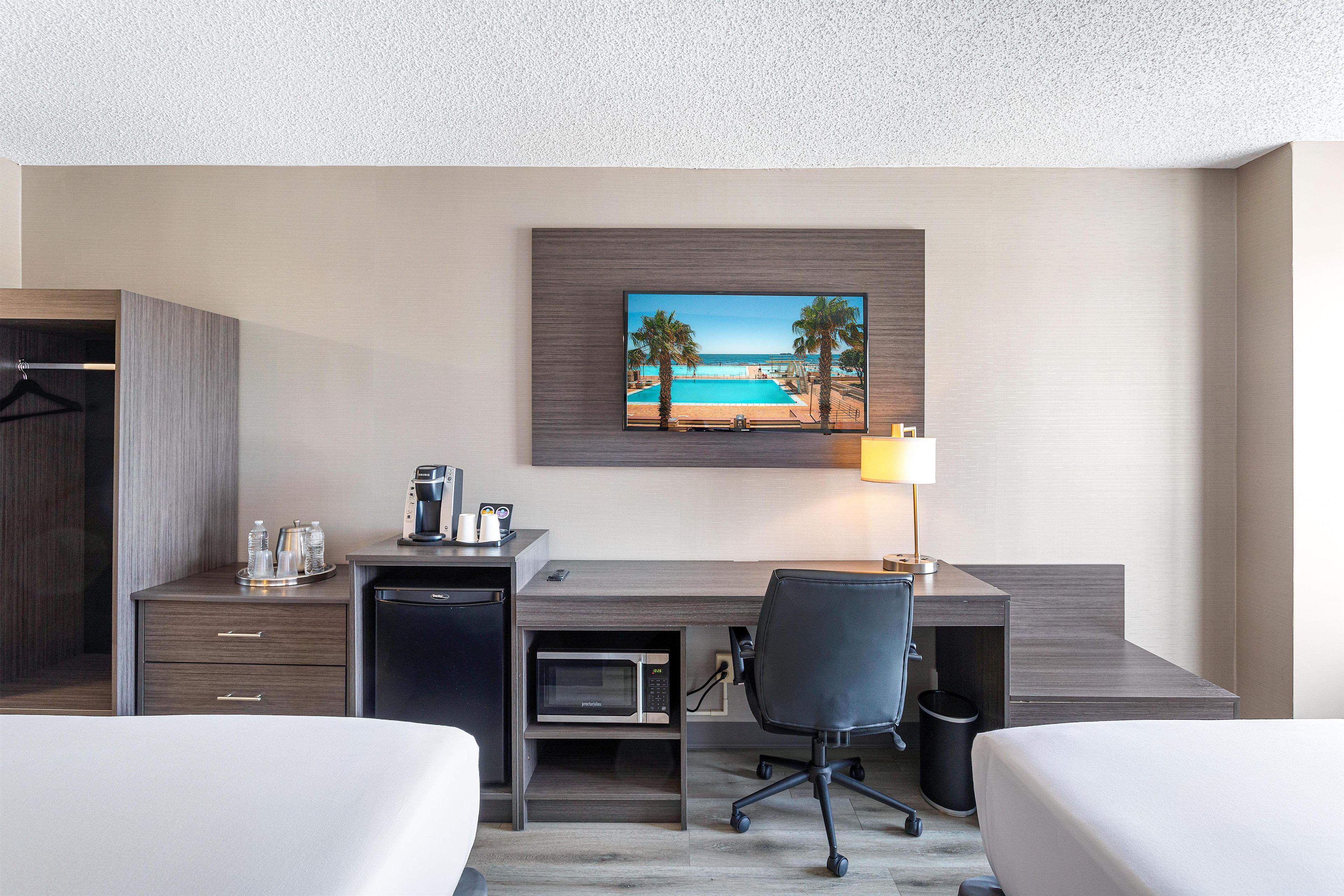 A hotel room with a desk, two beds, a coffee machine, a microwave, and a painting of a beach scene.