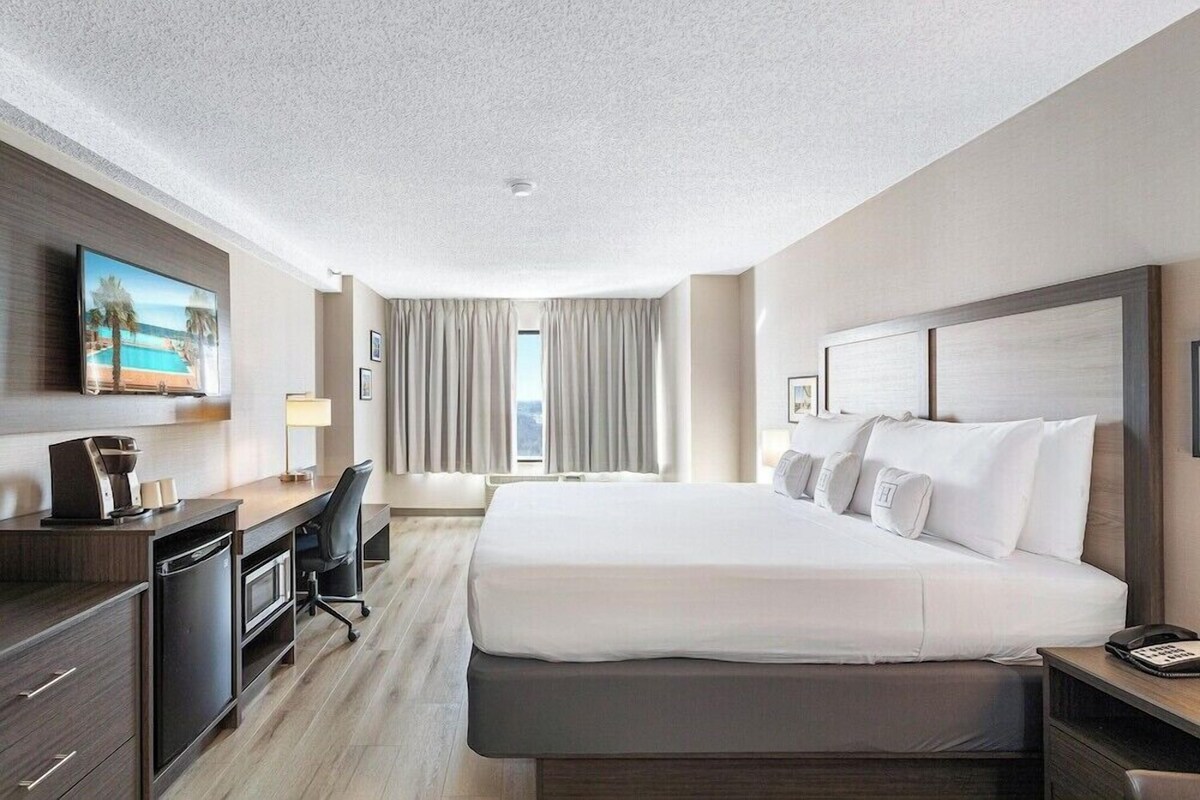 Habitación de hotel con una cama grande, un escritorio con ordenador, televisor y vistas al exterior.