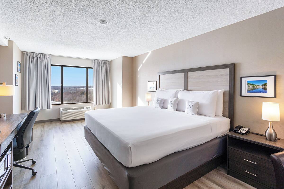 A hotel room with a large bed, a desk, and a chair. There is a window with curtains, a picture on the wall, and a lamp on the nightstand.