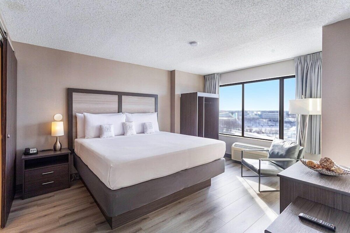 A modern hotel room with a large bed, a desk, a chair, a wardrobe, and a view of the city.