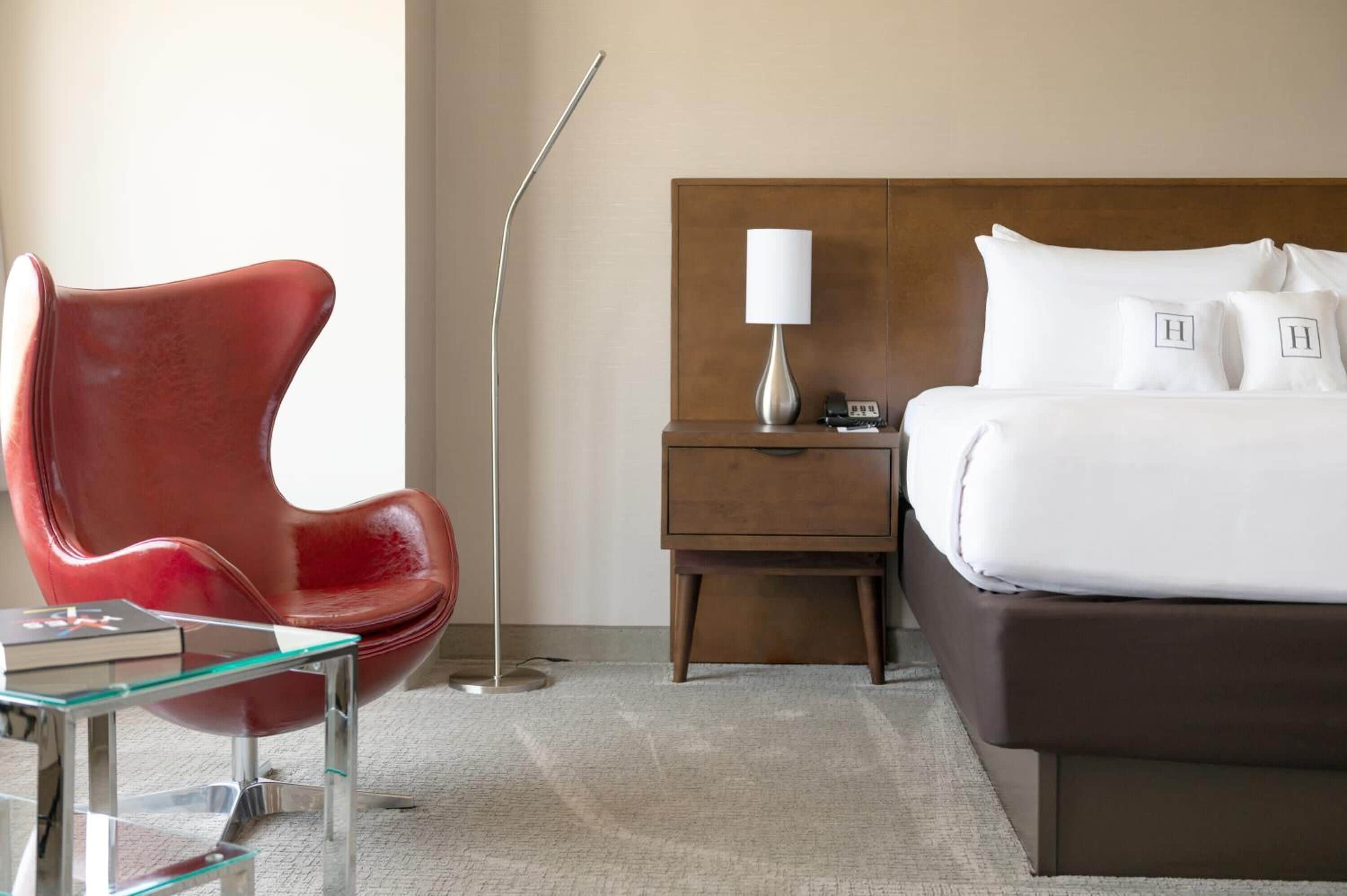 Camera d'albergo dal design moderno, con una poltrona in pelle rossa, un tavolino in vetro, un comodino in legno con una lampada e un letto grande con lenzuola e cuscini bianchi.