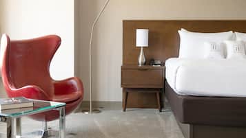 Camera d'albergo dal design moderno, con una poltrona in pelle rossa, un tavolino in vetro, un comodino in legno con una lampada e un letto grande con lenzuola e cuscini bianchi.