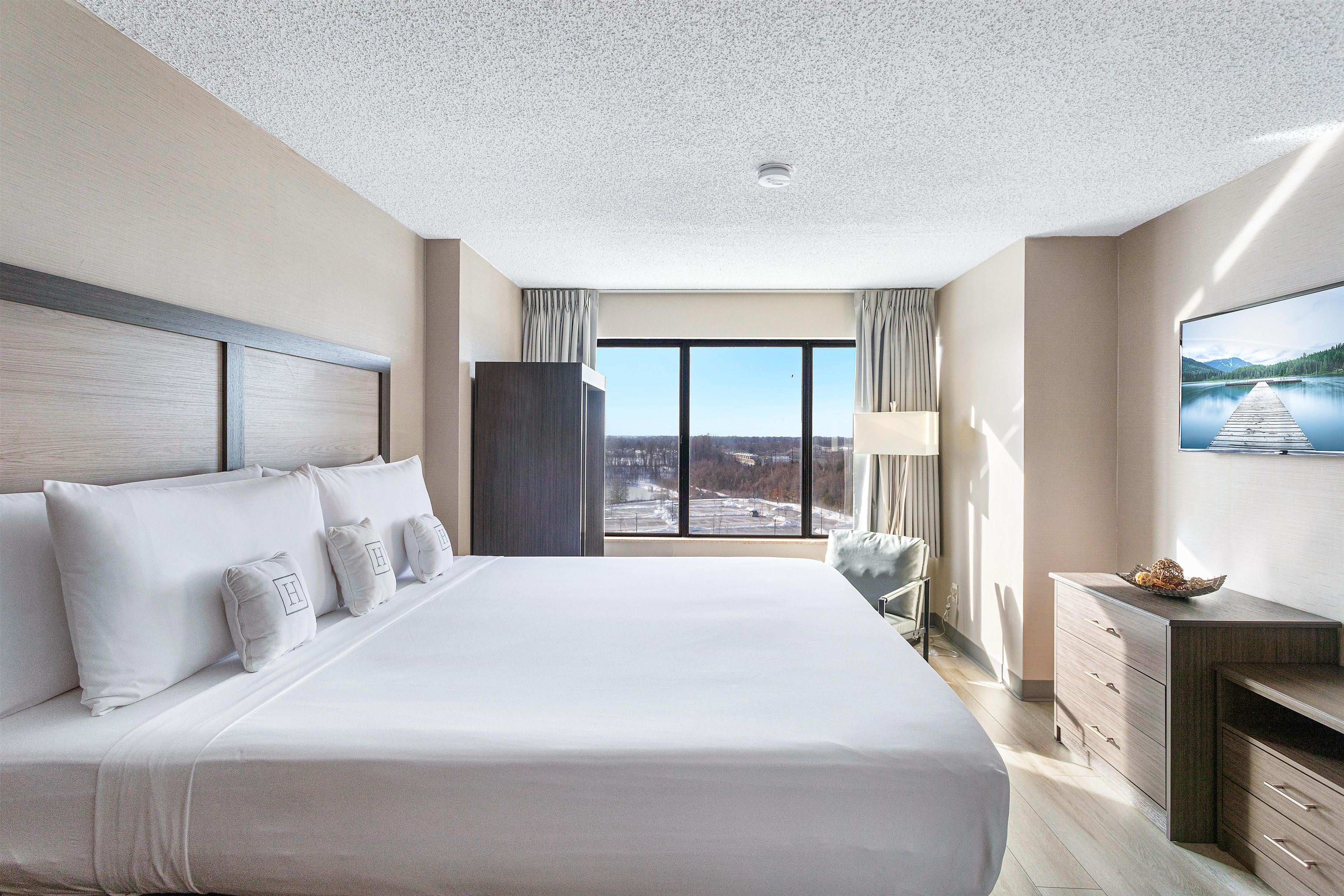 A hotel room with a large bed, a desk with drawers, a chair, a television, and a view of a lake through the window.