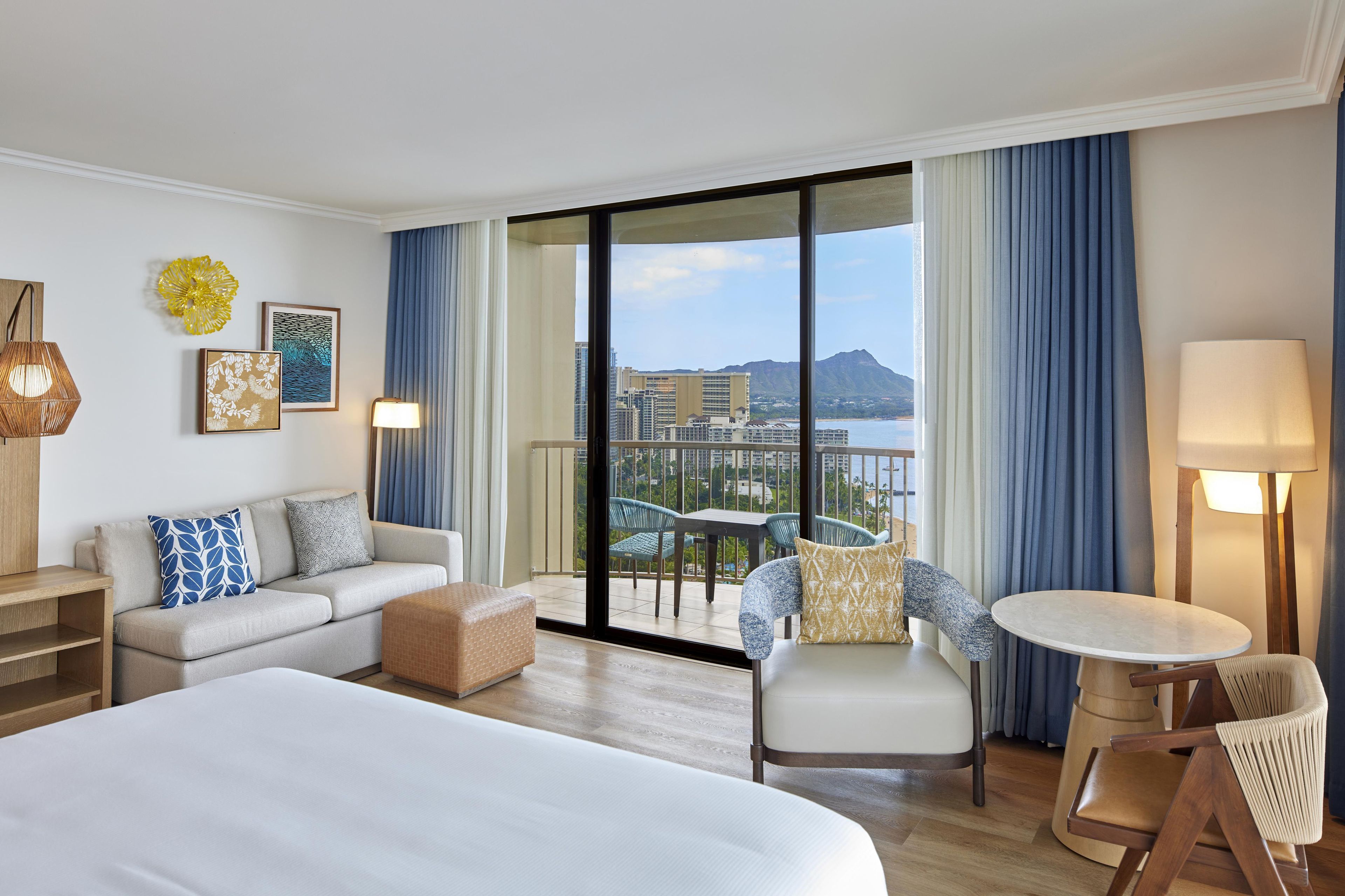 A hotel room with a large bed, a sofa, a chair, a small table, and a balcony with a view of buildings and a mountain.