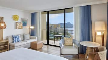 A hotel room with a large bed, a sofa, a chair, a small table, and a balcony with a view of buildings and a mountain.