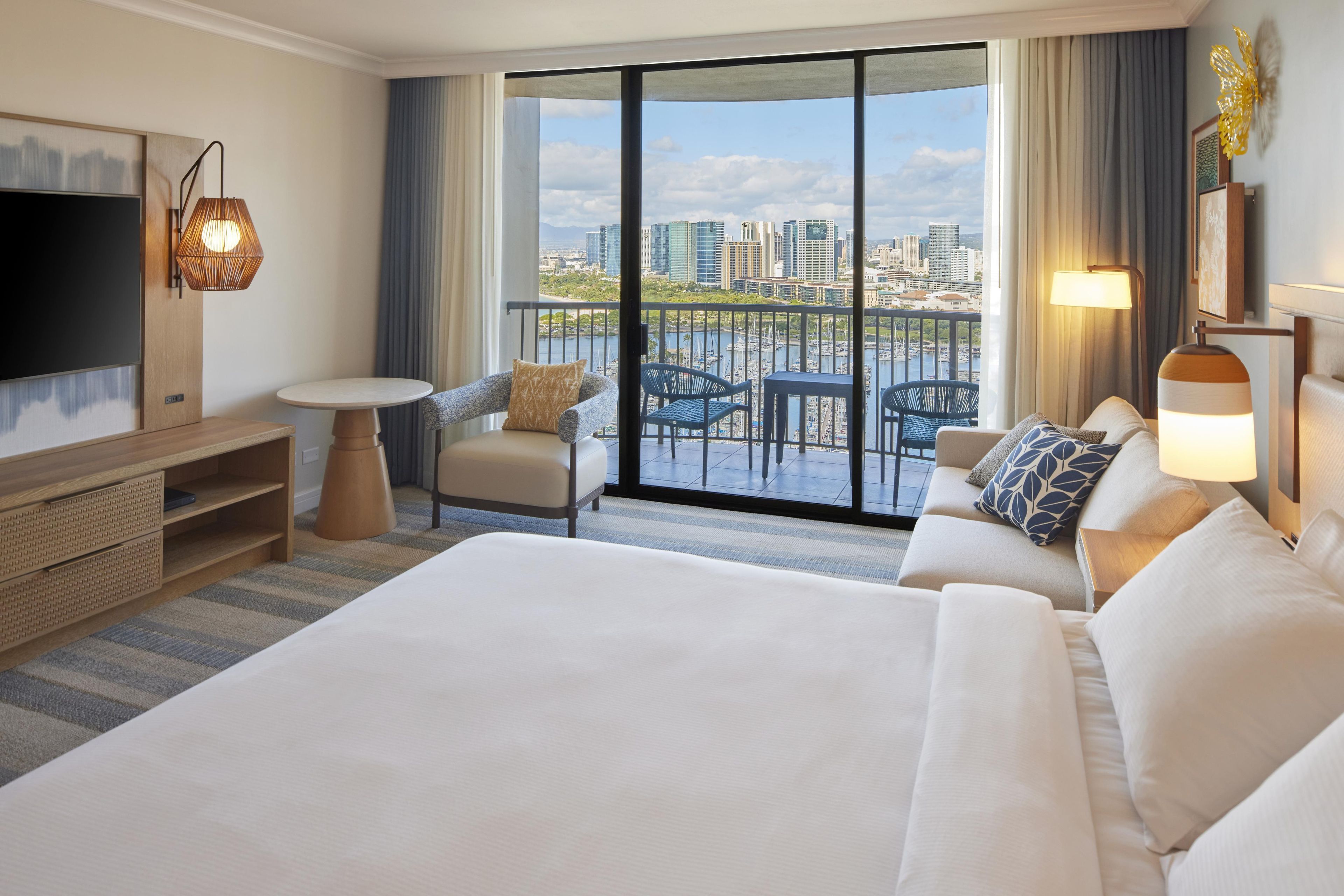 A hotel room with a large bed, a TV, a balcony with a city view, and a dining area.
