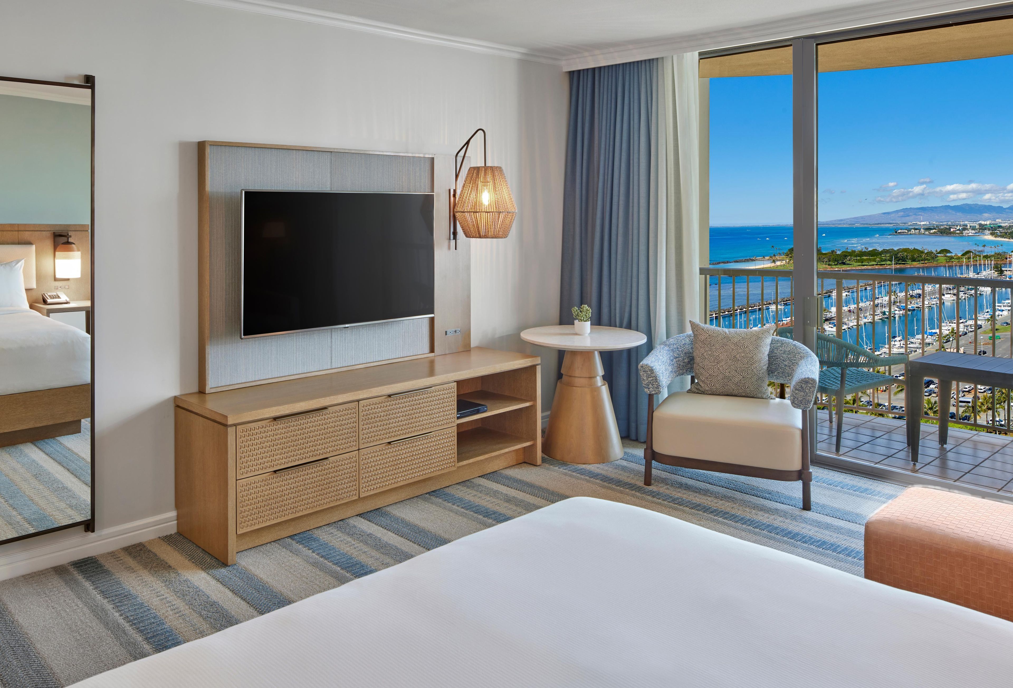 A hotel room with a large bed, a flat-screen TV, a wooden console, a round table, a chair, and a view of a marina and ocean.