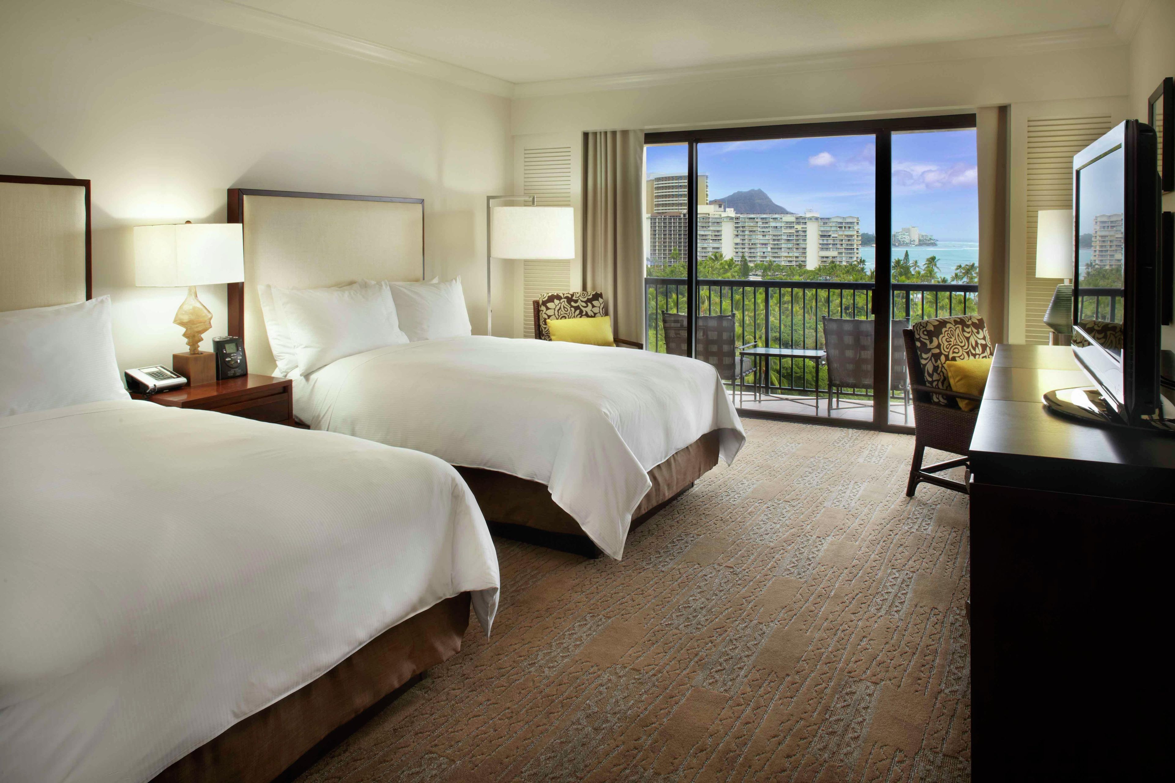 A hotel room with two beds, a desk, a chair, and a balcony with a view of buildings and the ocean.