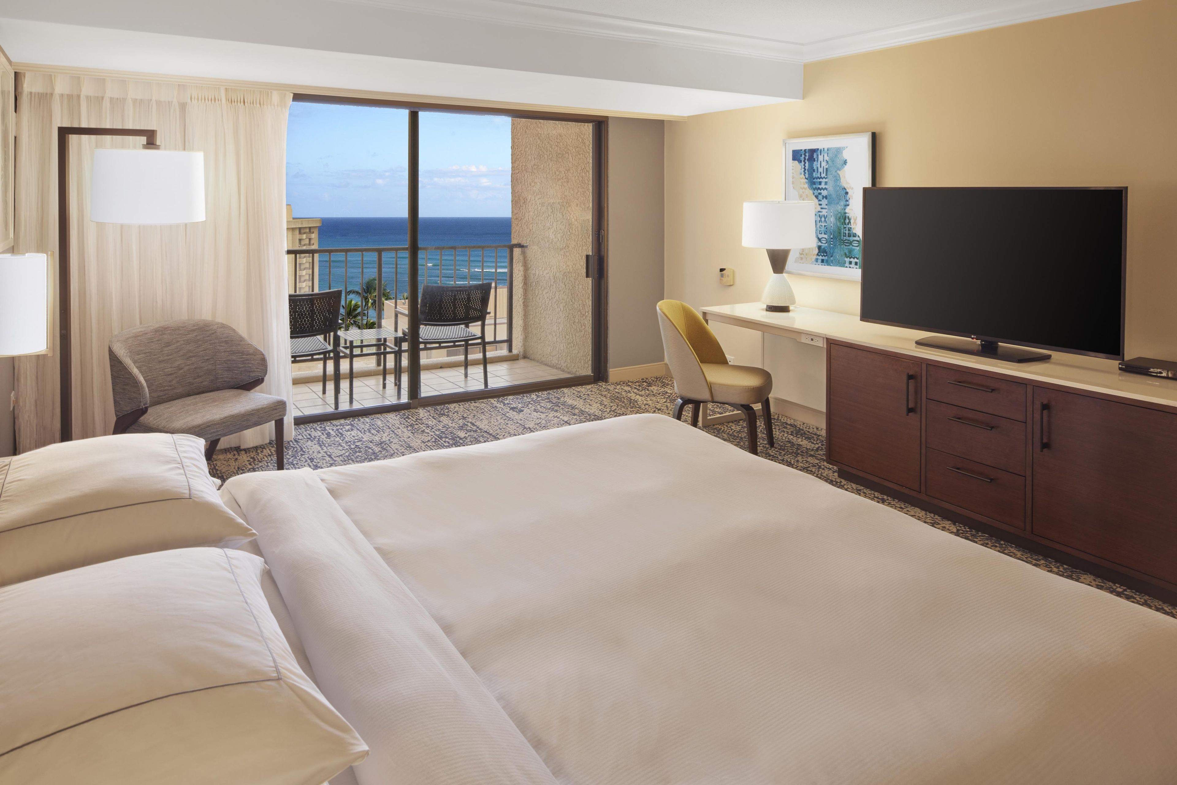 A hotel room with a large bed, a desk with a television, a chair, and a view of the ocean.