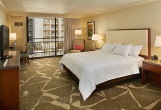 A hotel room with a large bed, a desk with a television, a chair, a balcony with a view of a building, and a painting on the wall.