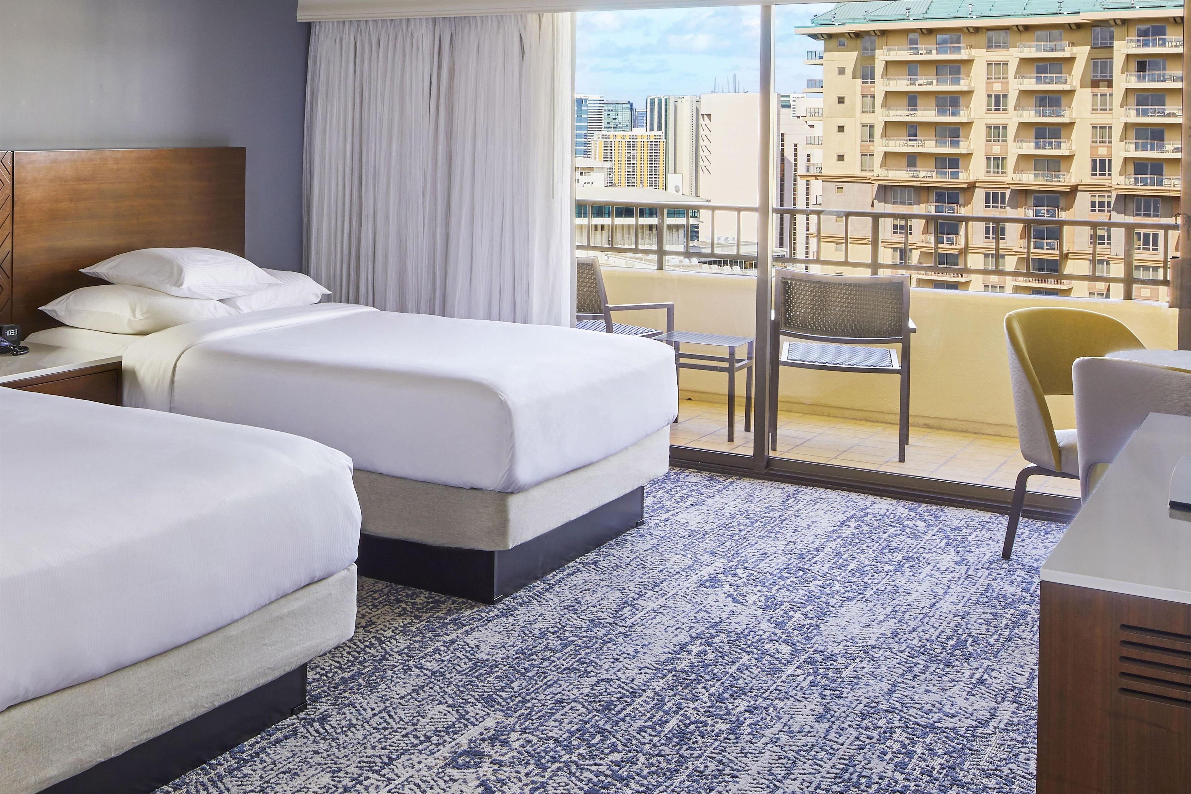 A hotel room with two beds, a nightstand, a chair, and a view of the cityscape through large windows.