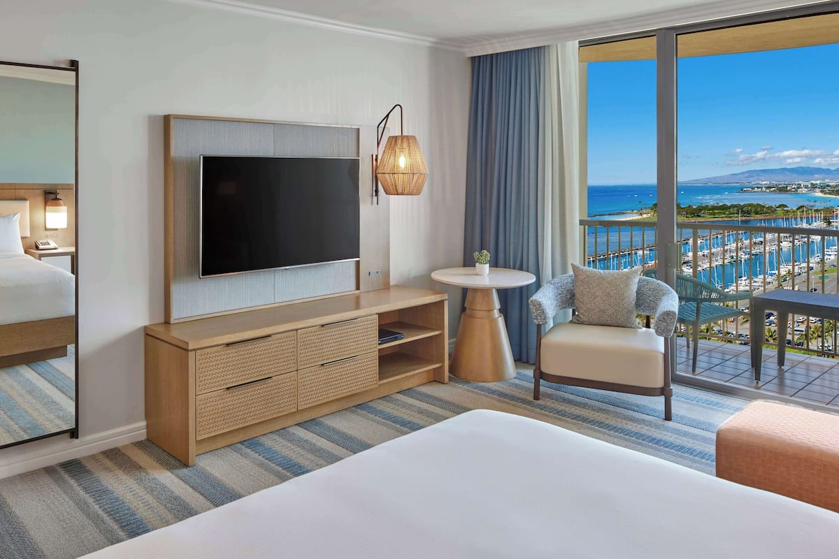 Una habitación de hotel con una cama grande, un televisor, un armario de madera, una mesa redonda con una planta y un balcón con vista al mar y barcos.