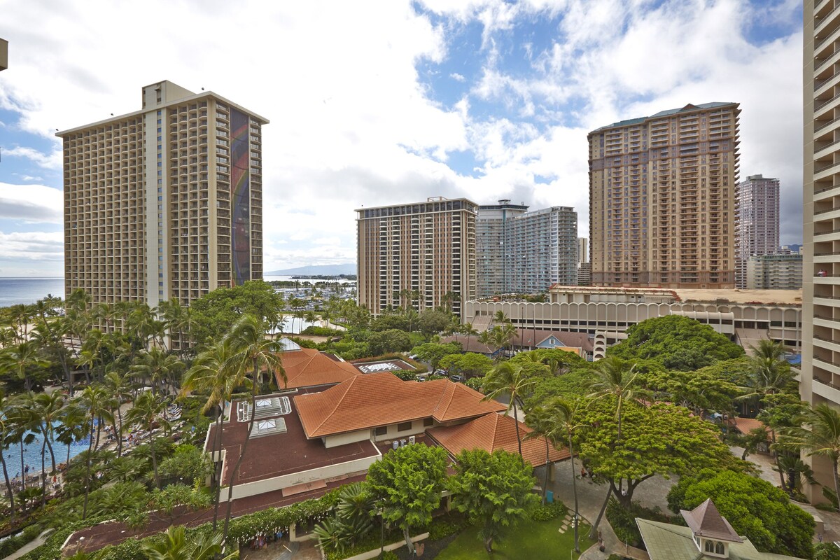 A high-rise hotel with multiple balconies, surrounded by lush greenery and a view of the ocean.