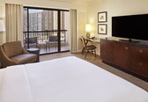 A hotel room with a large bed, a desk with a chair, a television, and a balcony with a view of high-rise buildings.