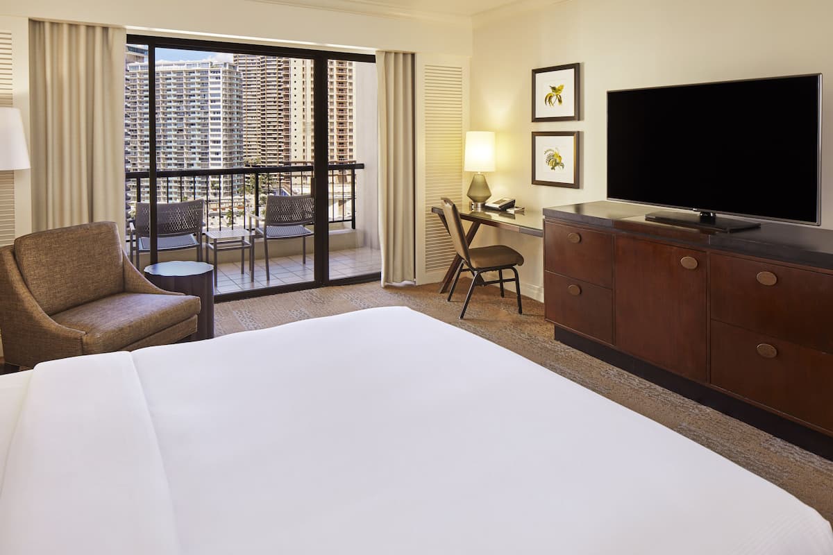 Una habitación de hotel con una cama grande, un escritorio con silla, televisión y un balcón con vista a edificios altos.