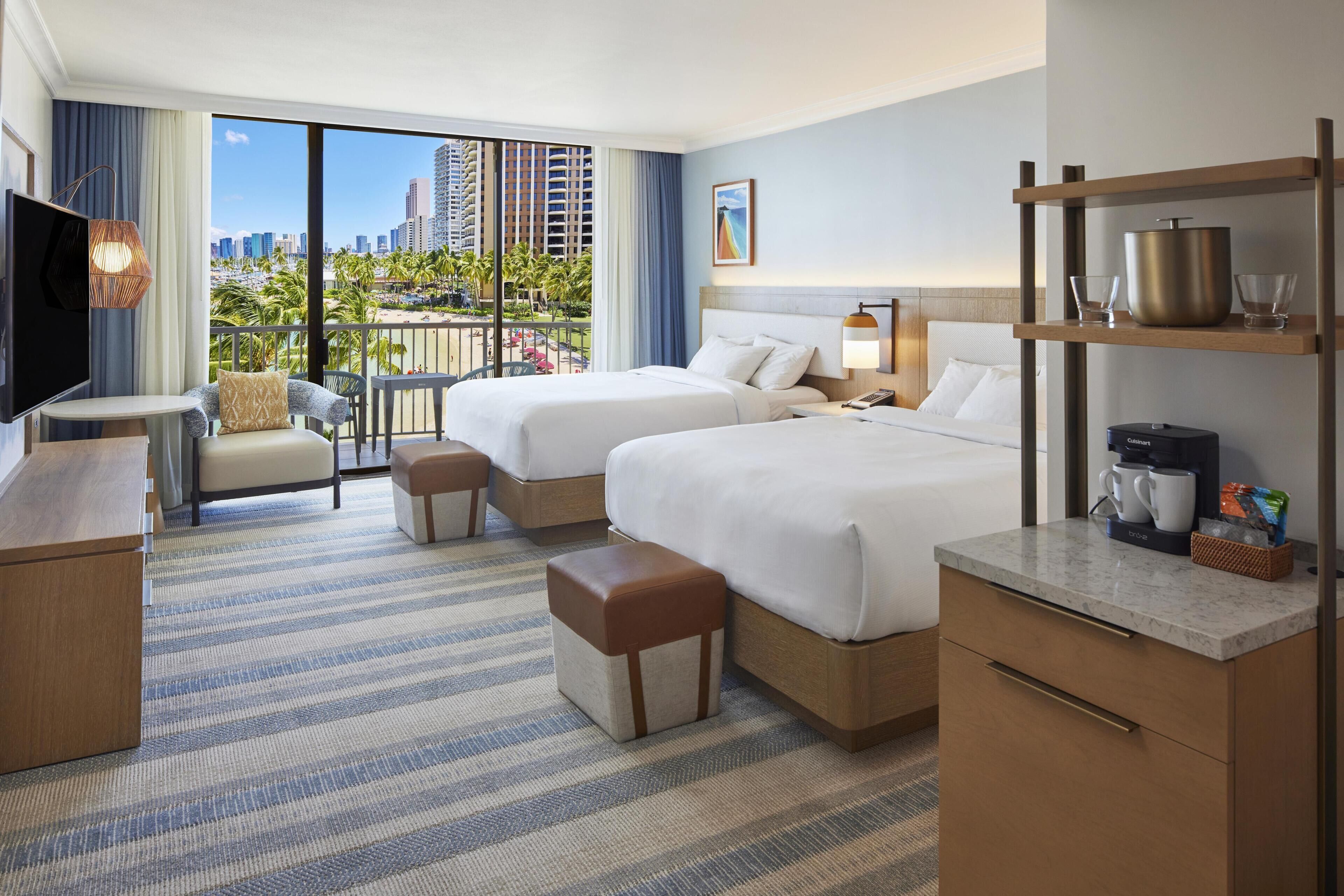 A hotel room with two beds, a TV, a desk, and a balcony with a city view.