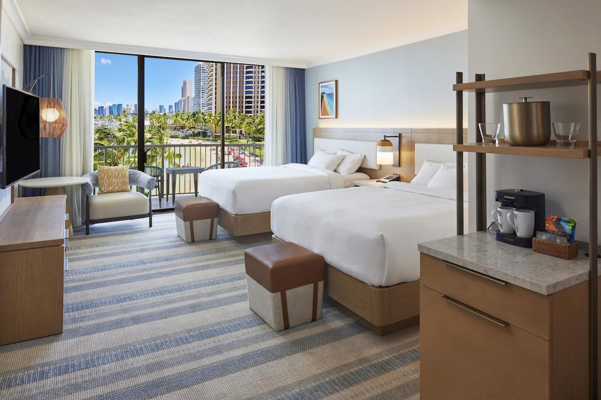 Habitación de hotel con dos camas, televisión, escritorio y balcón con vista a la ciudad.