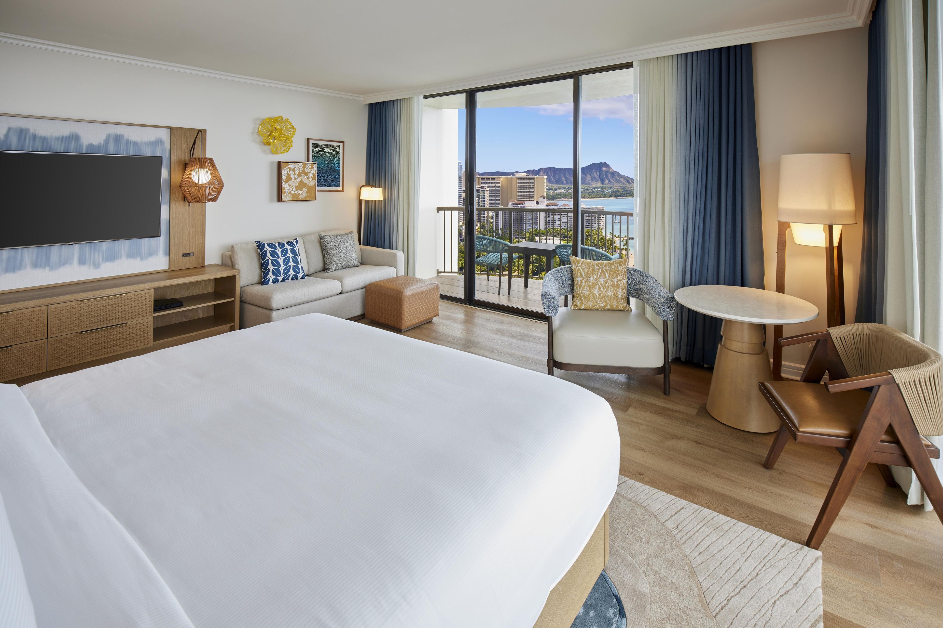 A hotel room with a large bed, a sofa, a small table, and a chair. There is a TV mounted on the wall and a balcony with a view of the city.