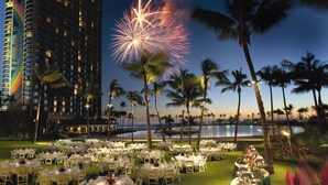 Un área de comedor al aire libre frente a la playa, con mesas preparadas para un evento formal, con vista a una masa de agua y un alto edificio de hotel.