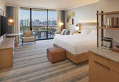 A hotel room with a large bed, a desk, a chair, a small table, and a balcony with a city view.