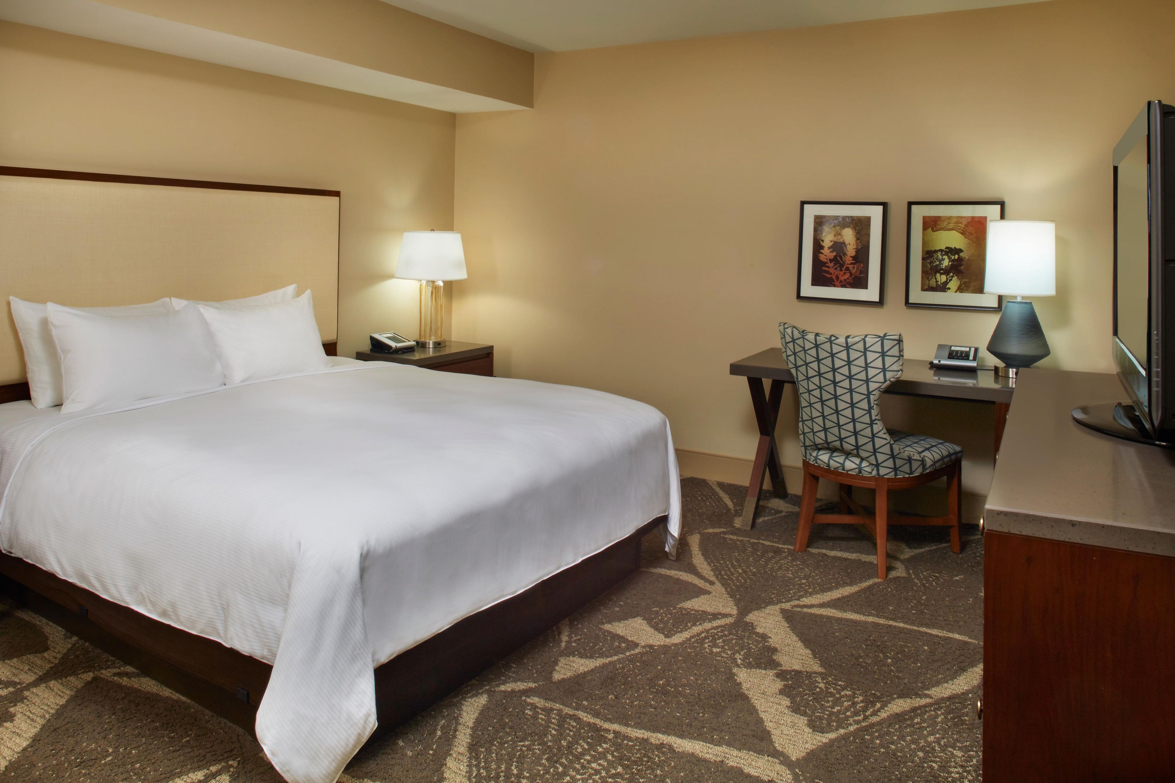 A hotel room with a large bed, a desk with a chair, two wall-mounted lamps, and two framed pictures.