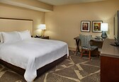 A hotel room with a large bed, a desk with a chair, two wall-mounted lamps, and two framed pictures.