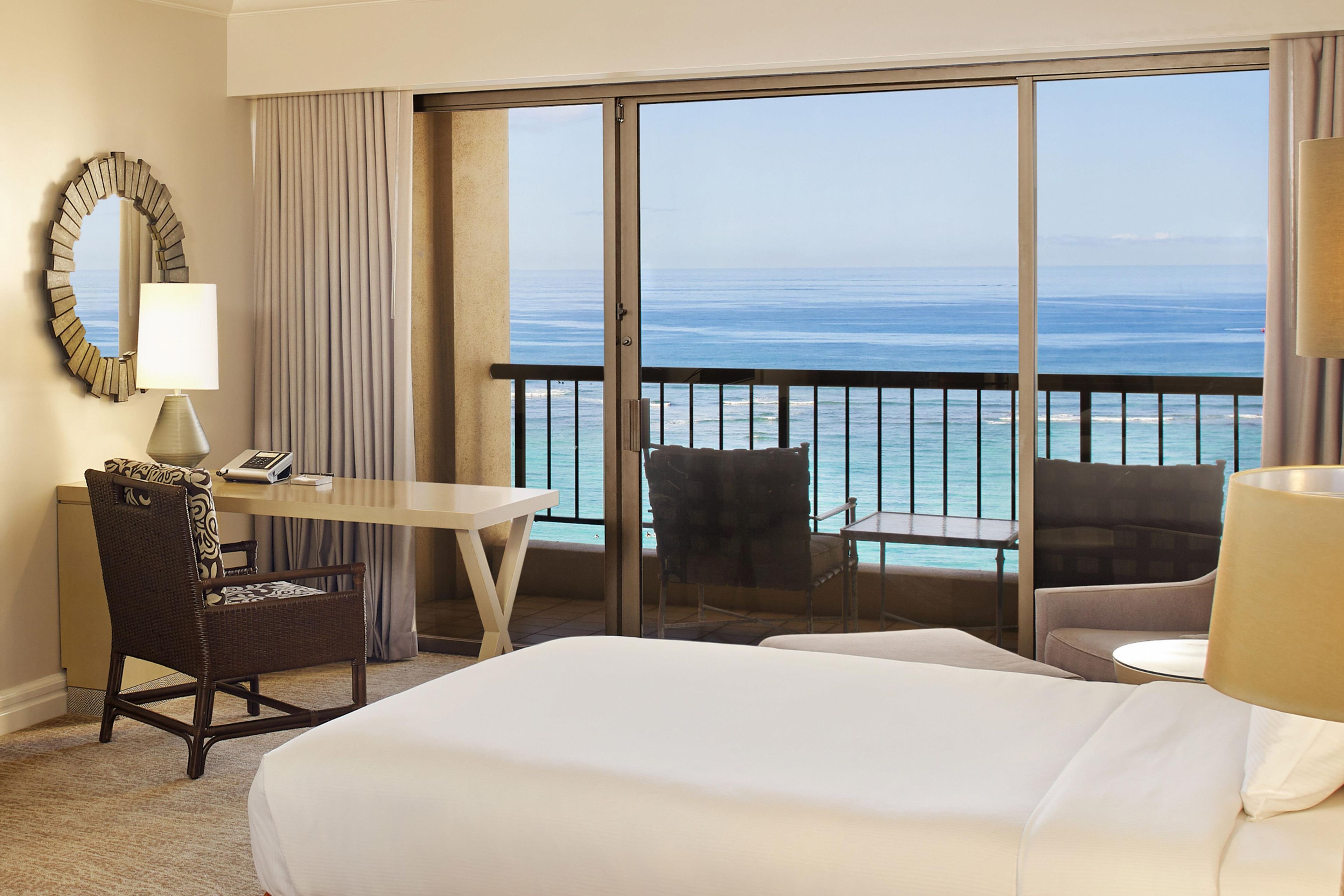 Une chambre d’hôtel avec un grand lit, un bureau, deux chaises, une lampe de bureau, un téléphone et une vue sur l’océan depuis un balcon.
