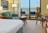 A hotel room with a bed, a green armchair, a round wicker table, and two chairs. A balcony with a view of the ocean and buildings is visible.