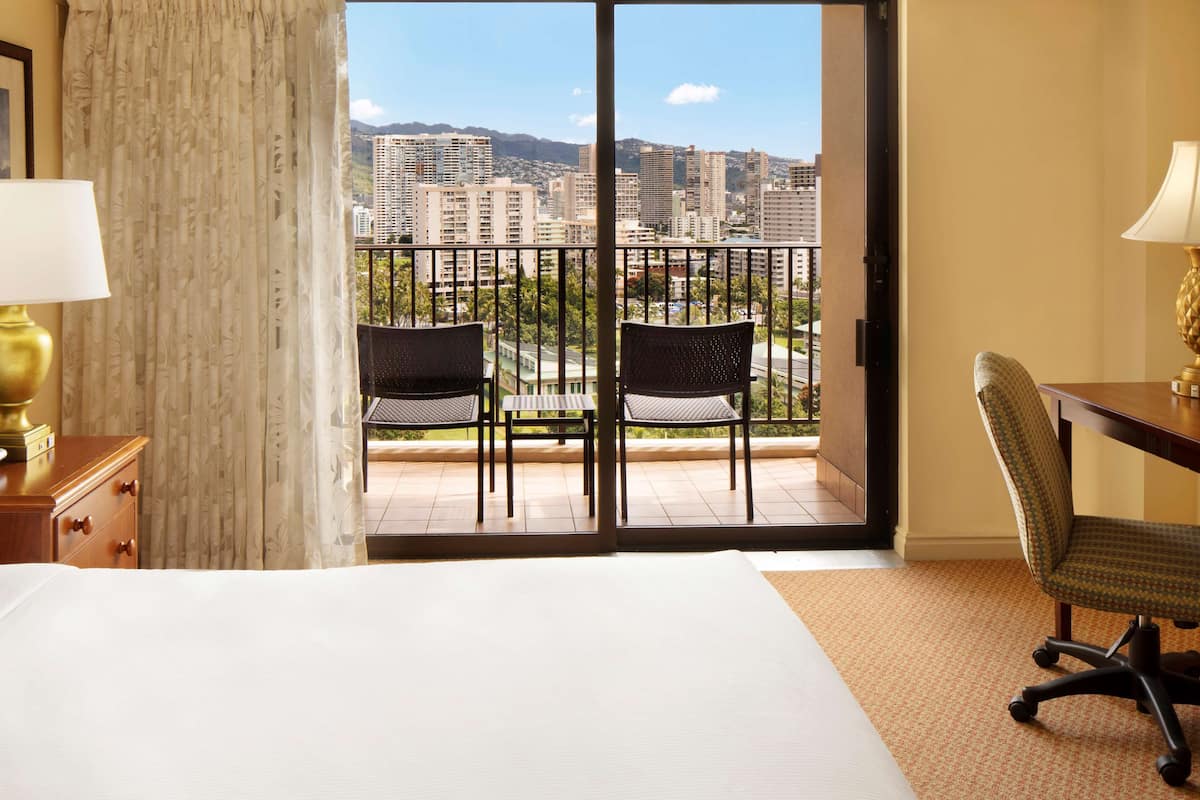 Habitación de hotel con cama, escritorio, dos sillas, vista de la ciudad a través de una puerta de balcón y una lámpara en el escritorio.