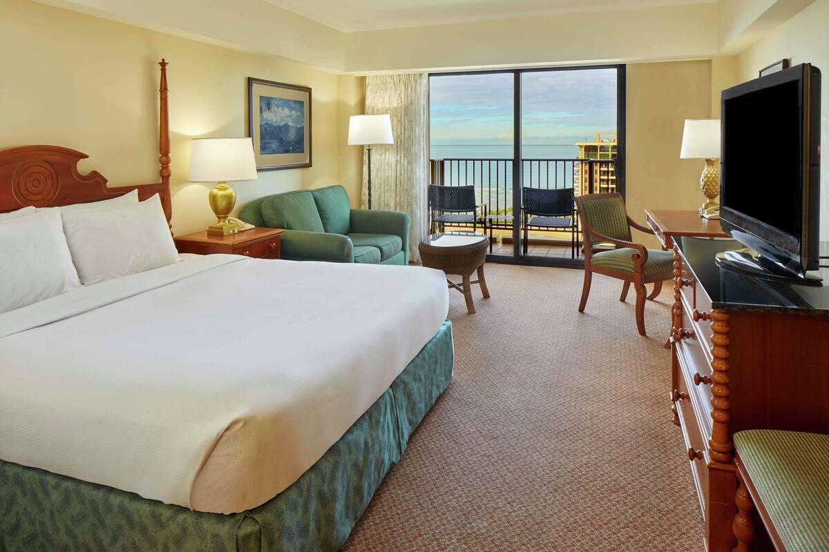 Habitación de hotel con una cama grande, televisión, balcón con vista al océano y una zona de estar con sillas.