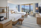 A hotel room with two beds, a desk, a coffee maker, a TV, and a view of the sea.