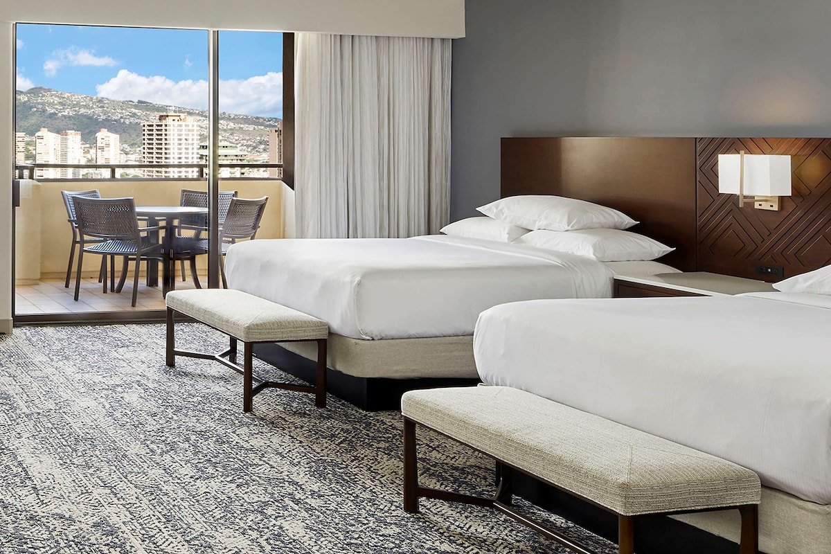 Habitación de hotel con dos camas, un balcón con mesa y sillas, y vistas a edificios y montañas.