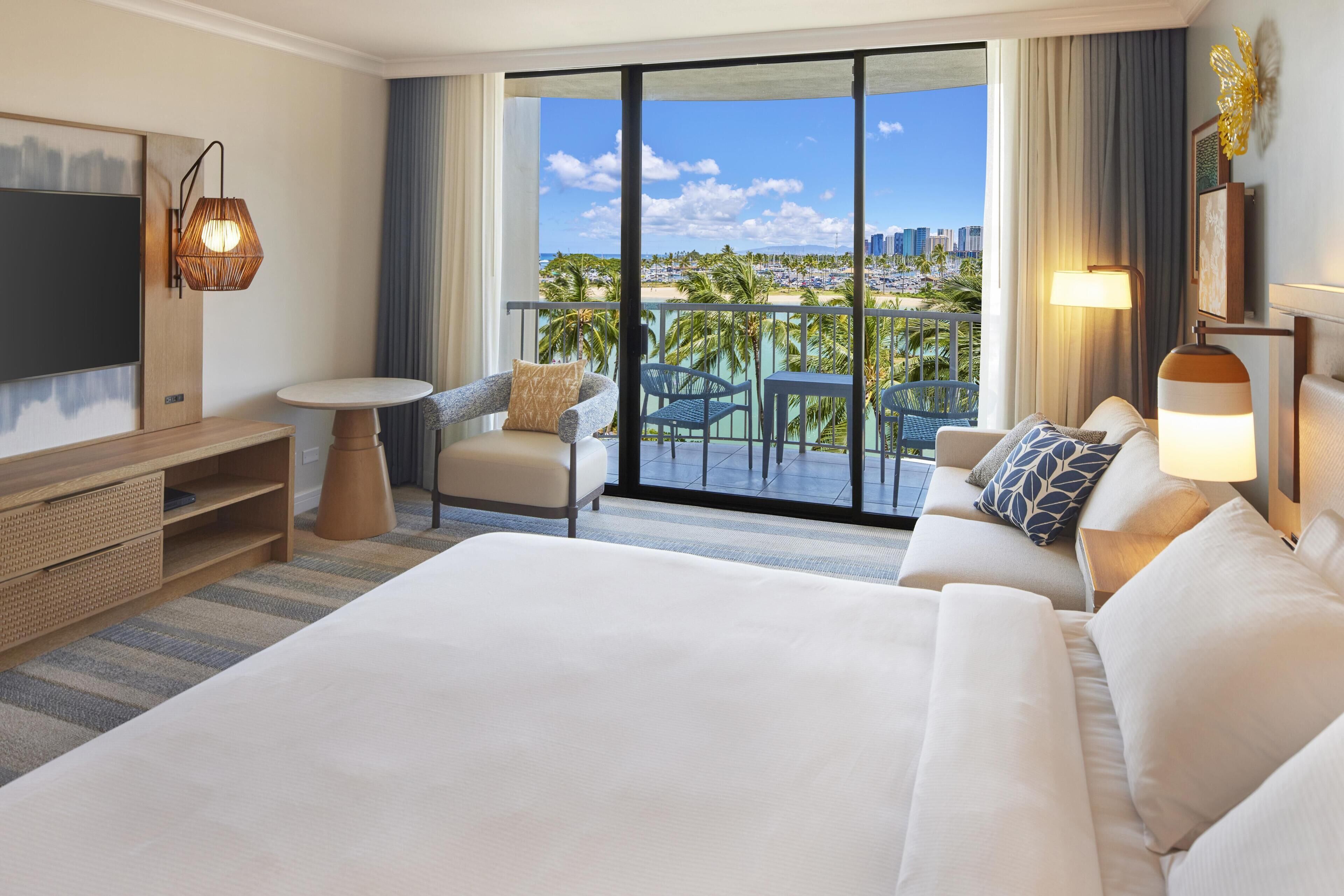 A hotel room with a large bed, a TV, a balcony with a city view, and a dining area.