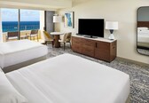 A hotel room with two beds, a TV, a desk with a chair, a dresser, and a large window with a view of the ocean.