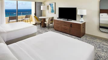 Habitación de hotel con dos camas, televisión, escritorio con silla, cómoda y un gran ventanal con vista al océano.