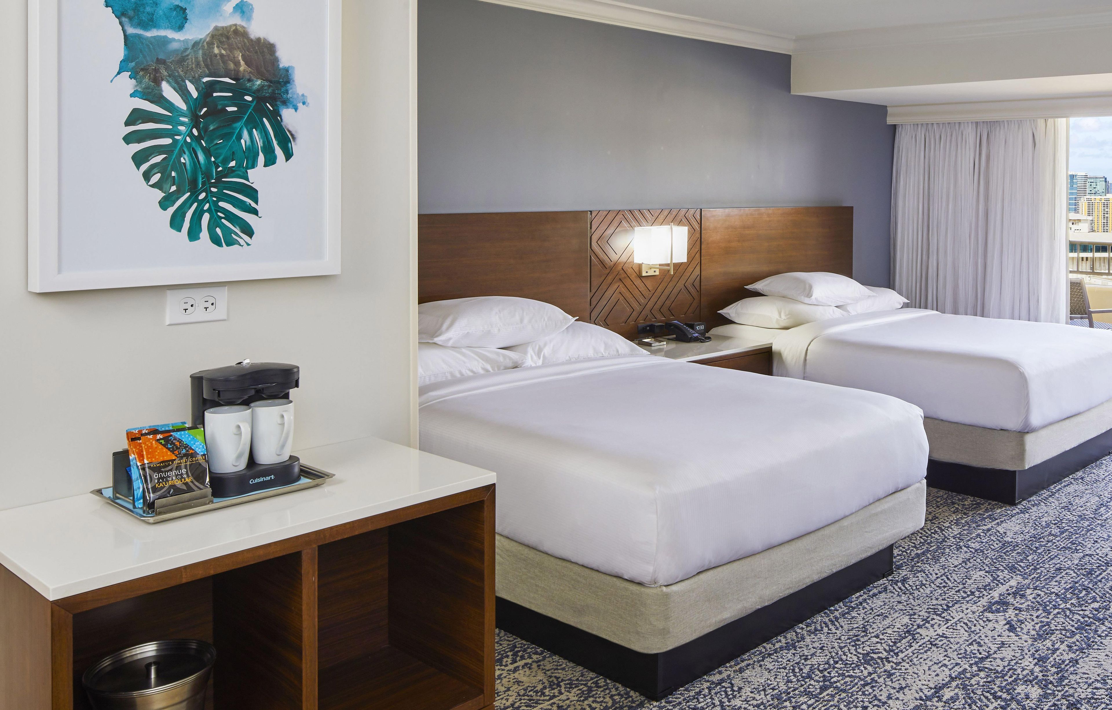 A hotel room with two beds, a nightstand with a coffee maker, a framed artwork, and a view of the city.