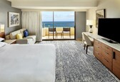 A hotel room with a large bed, a desk with a TV, a balcony with chairs, and a view of the sea.