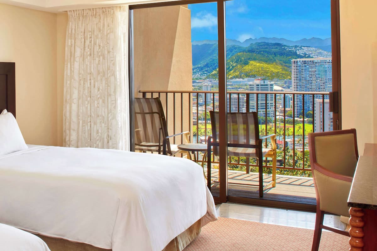 Habitación de hotel con cama, una silla, una mesa y balcón con vistas a la ciudad y a las montañas.