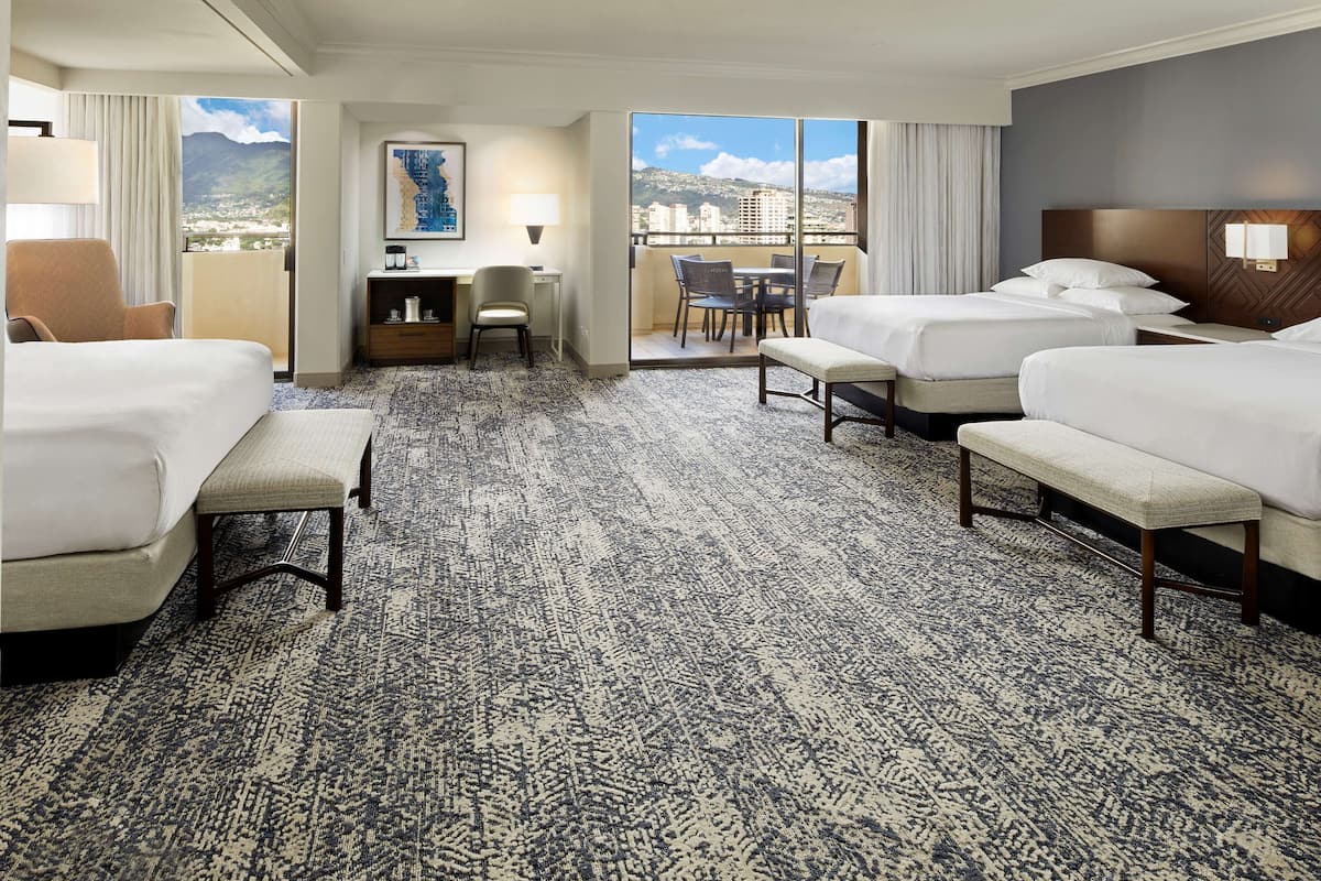 Habitación de hotel con tres camas, un escritorio y un balcón que ofrece vistas a la ciudad y a las montañas.