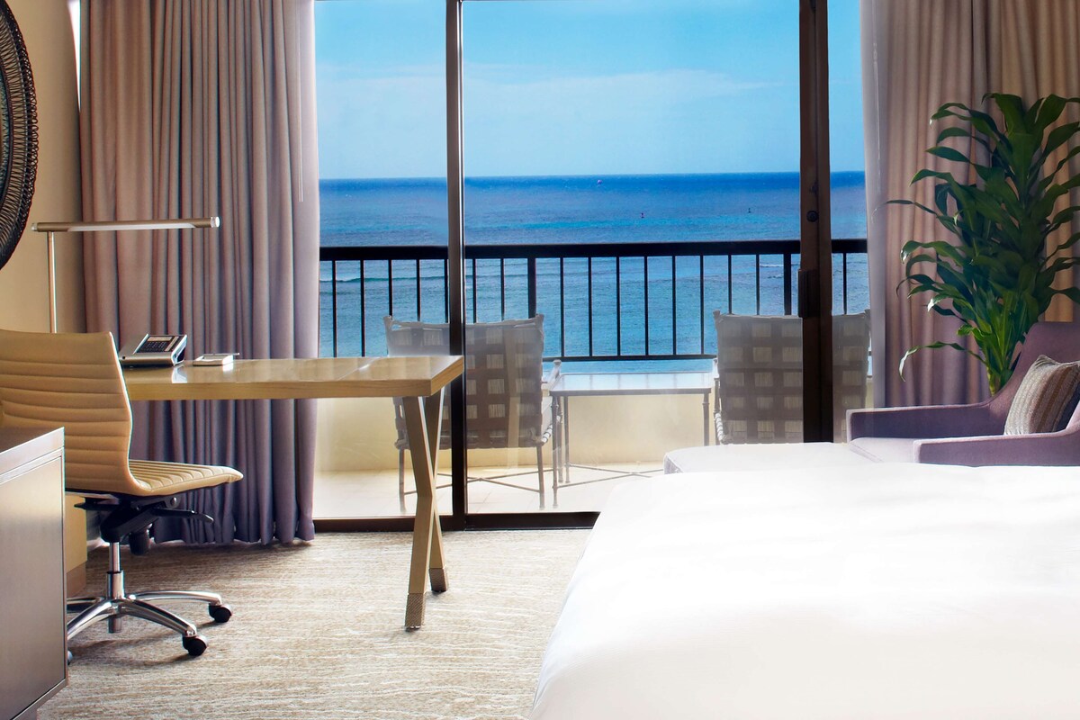 Habitación de hotel con una cama grande, un escritorio con silla, una mesita y vistas al océano a través de amplios ventanales.