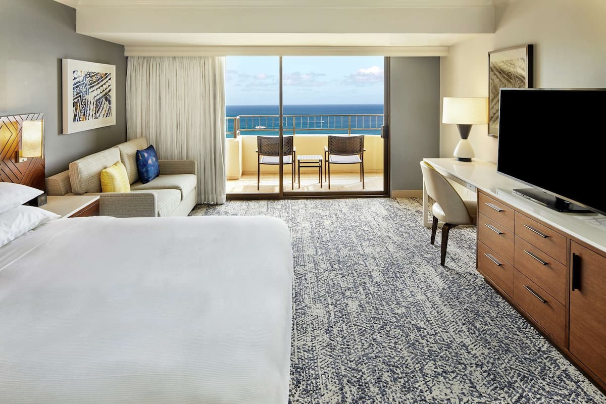 Habitación de hotel con una cama grande, un escritorio con televisor, un balcón con sillas y vista al océano.