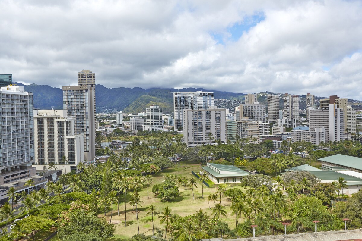 A cityscape with high-rise buildings, a park with palm trees, and mountains in the background.