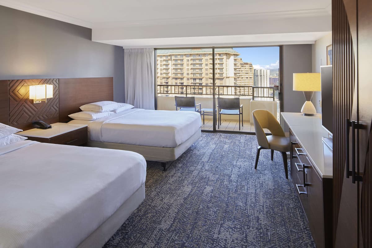 Habitación de hotel con dos camas, un escritorio, una silla y un balcón con vistas a la ciudad.