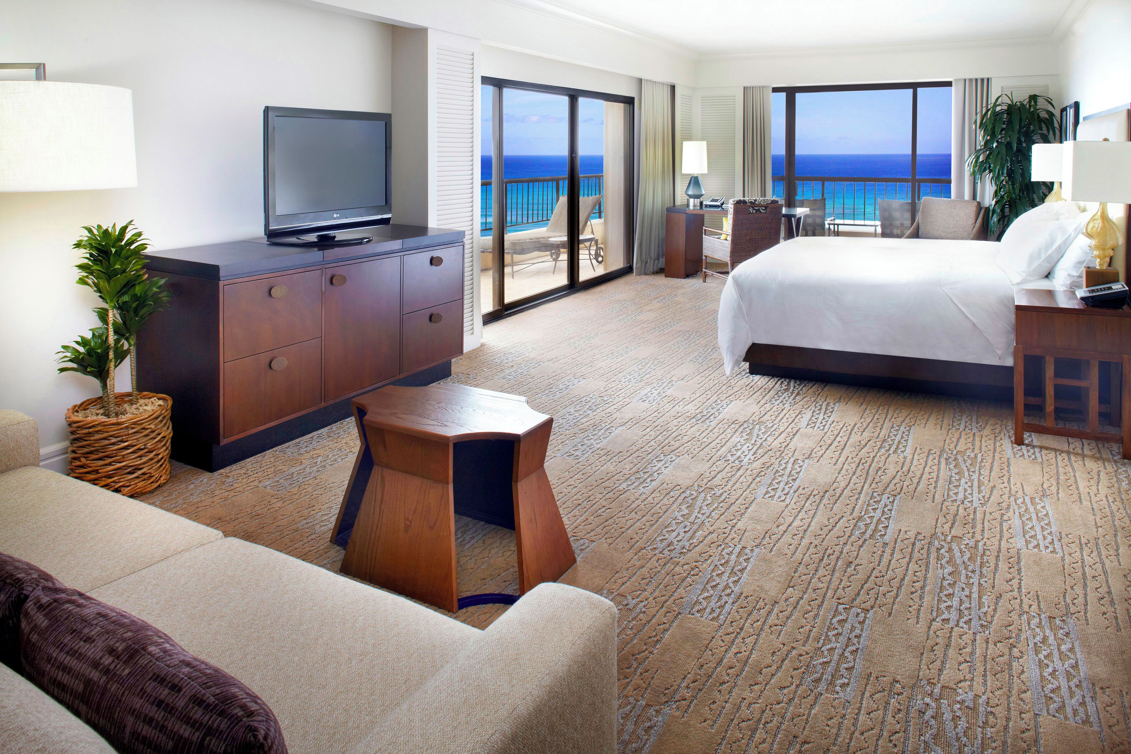 Habitación de hotel con una cama grande, un televisor sobre un mueble de madera, una mesita pequeña, un sofá y vista al océano a través de la puerta corrediza de vidrio.