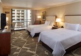 A hotel room with two beds, a TV, a desk, a chair, a balcony, and a view of a building.