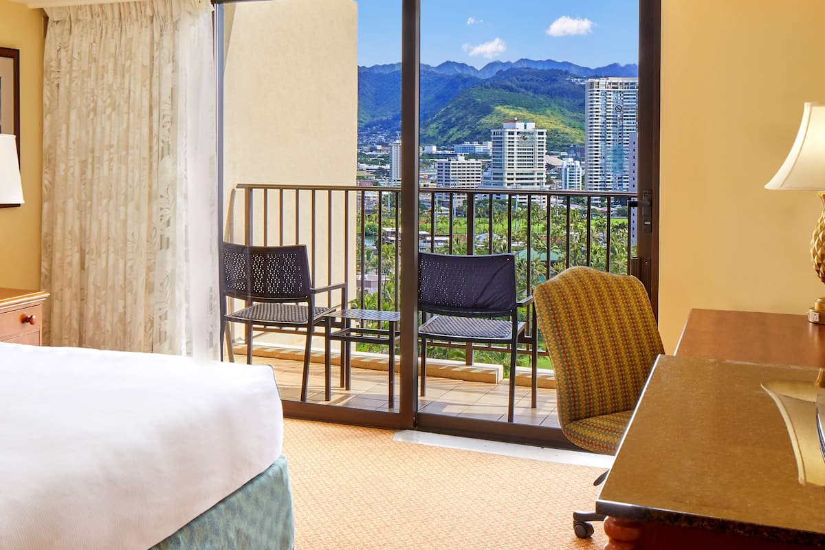 Habitación de hotel con cama, escritorio y vistas a un paisaje urbano y montañas.